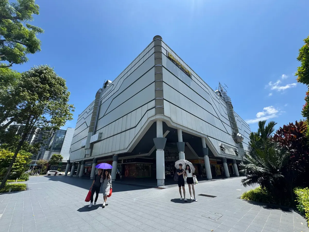 Yishun 10 at street level: the white four-storey cinema block on pillars, with shoppers walking across the paved plaza in front on a sunny day